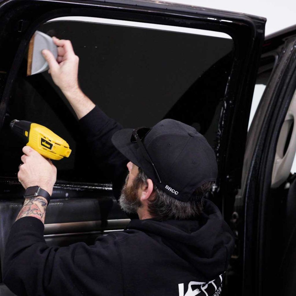 A person in a black hoodie and cap applies window tint to a car door using a heat gun and a squeegee.
