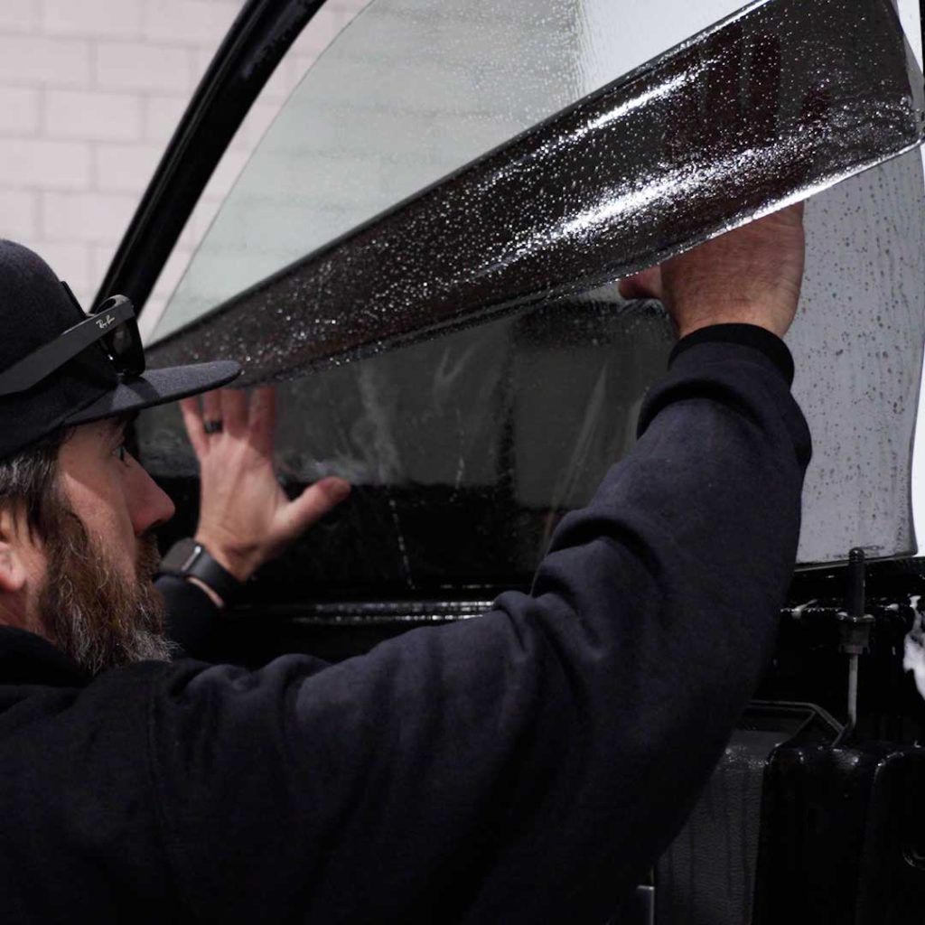 A person in a black cap and hoodie applies a layer of wet window tint film to a car door glass.