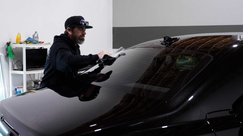 A person wearing a black hoodie and cap uses a tool to apply window tint to the rear windshield of a black car.
