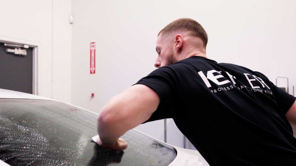A person in a black t-shirt wipes soapy water off the rear windshield of a vehicle in an indoor shop.