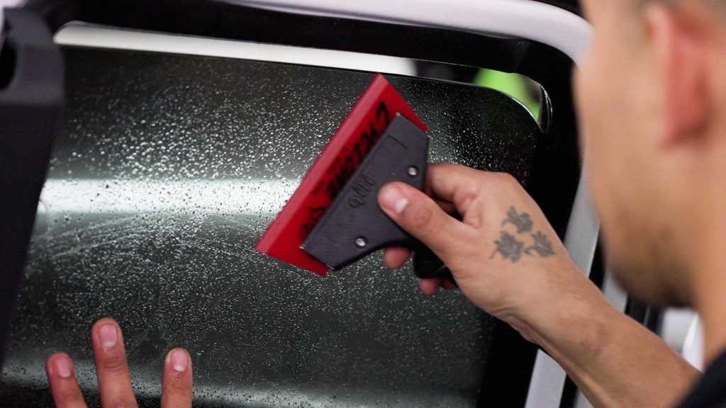 A hand uses a red squeegee to smooth window tint film onto a car window.