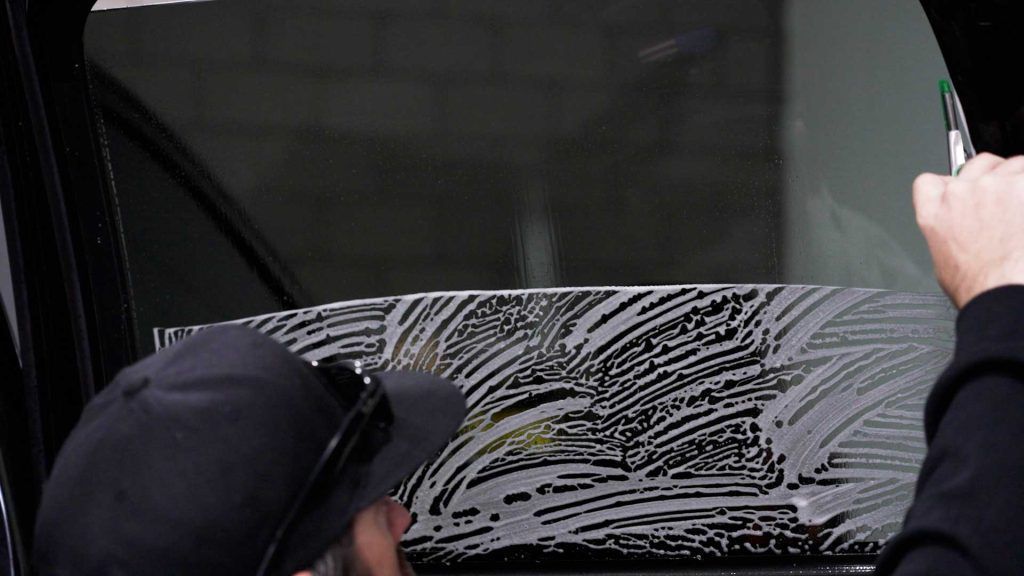 A person in a black cap cleans a vehicle window with a soapy, streaked cleaning solution.